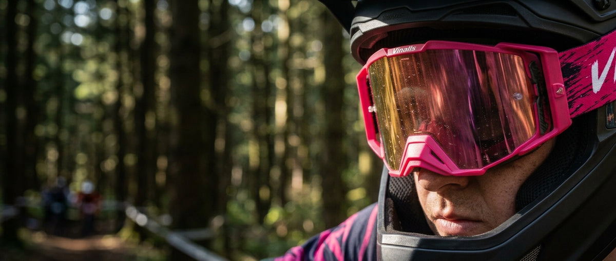 Close-up of a person wearing bright pink ski goggles with a reflective gold-orange lens, mounted on a black helmet, set against a blurred forest background.